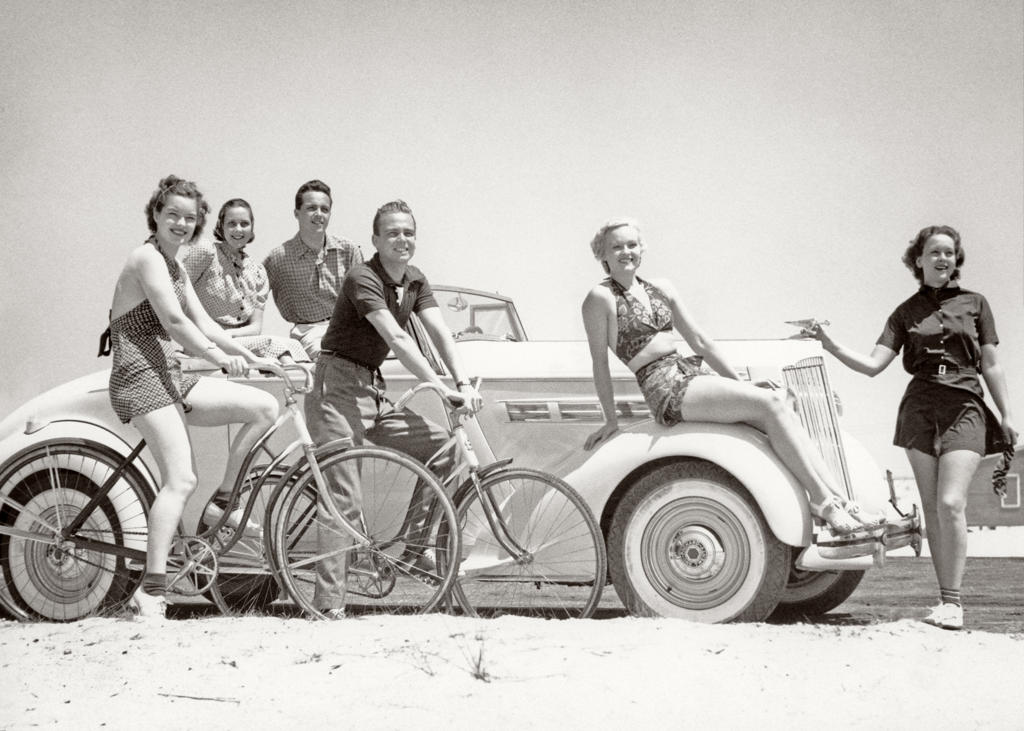 1930s GROUP OF MEN AND WOMEN WEARING BATHING SUITS CASUAL CLOTHES ON BICYCLES IN A CAR ON BEACH ALL LOOKING AT CAMERA SMILING