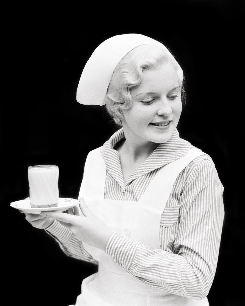 1930s PRETTY BLONDE NURSE HOLDING A DISH WITH GLASS OF MILK LOOKING DOWN TO TALK TO A PATIENT