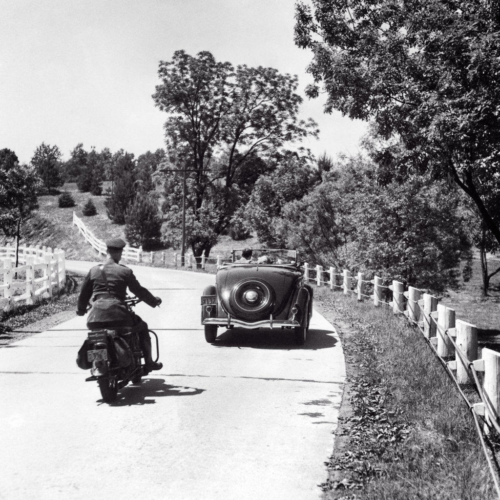1930s 1940s MAN HIGHWAY PATROL POLICE OFFICER ON MOTORCYCLE PURSUING CONVERTIBLE CAR WITH COUPLE TWO PASSENGERS ON COUNTRY ROAD