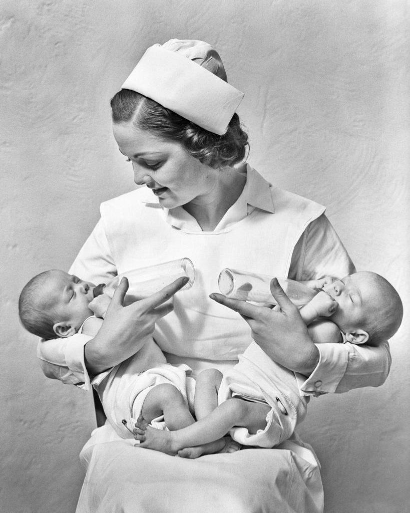 1930s SMILING NURSE SITTING HOLDING JUGGLING TWO NEWBORN TWIN BABY GIRL SISTERS IN HER ARMS FEEDING THEM WITH BOTTLES