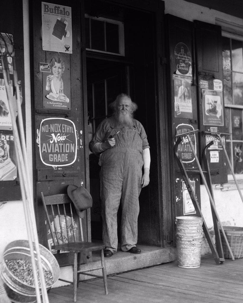 1930S Elderly Bearded Man In Overalls Smoking Pipe Standing In Doorway Of General Store