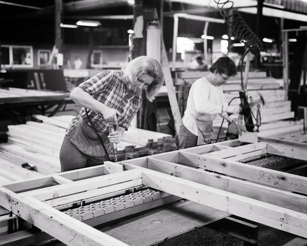 1960s 1970s TWO WORKERS WOMEN ON PREFAB FACTORY FLOOR ASSEMBLING A WOODEN FRAME FOR USE IN PREFABRICATED BUILDING 
