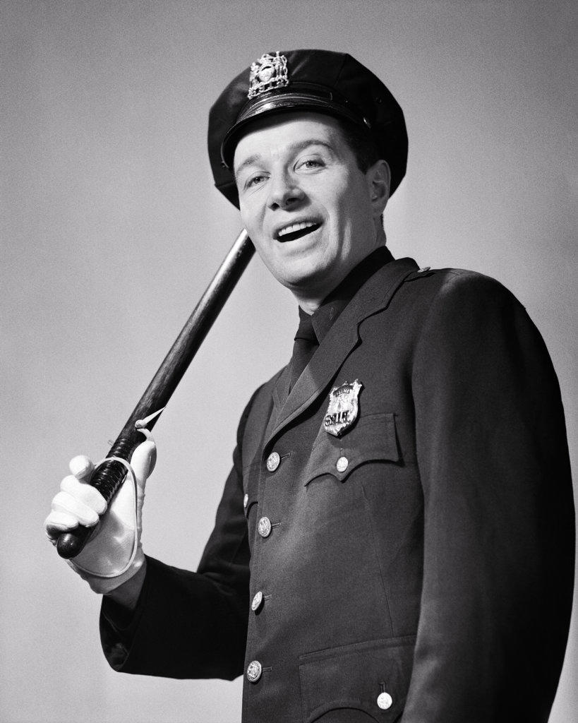 1950s SMILING UNIFORMED BEAT POLICEMAN LOOKING AT CAMERA TIPPING HIS HAT WITH NIGHT STICK BILLY CLUB