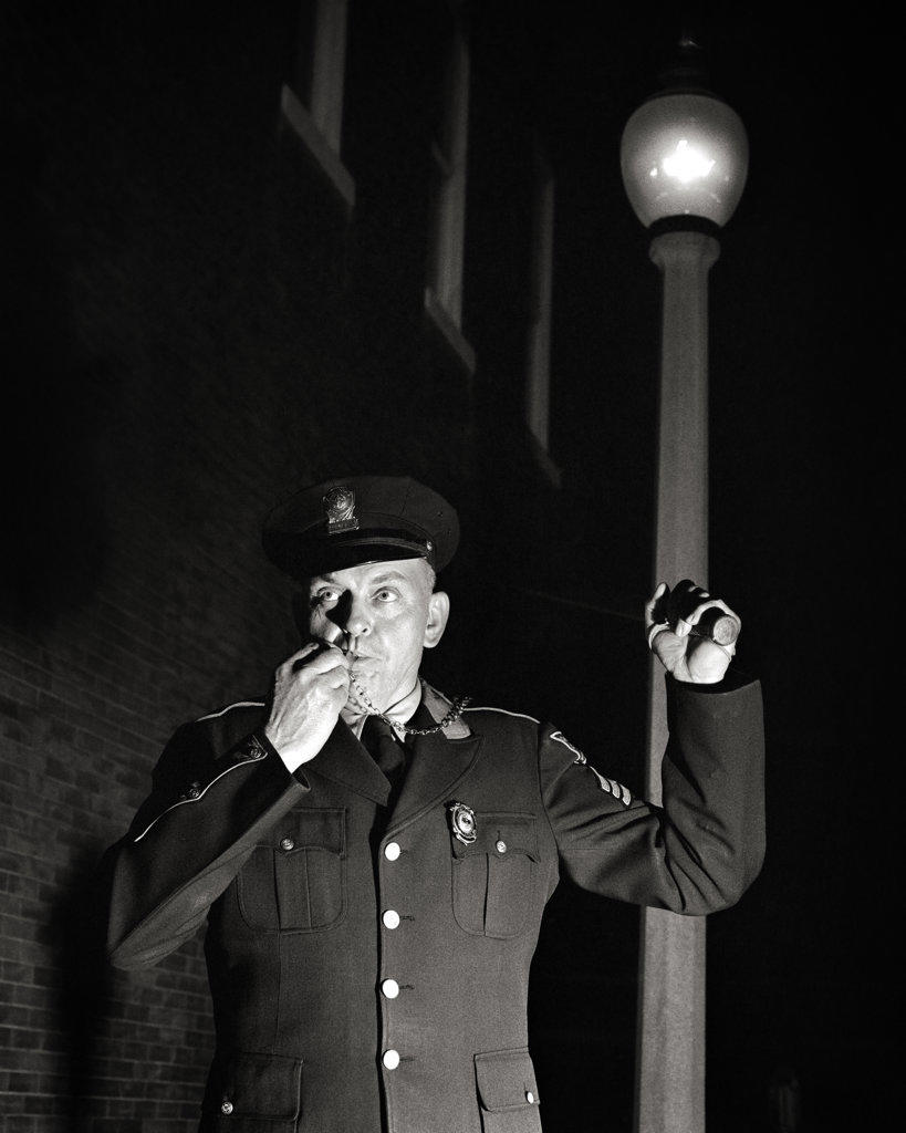 1950s SINGLE FOOT PATROL POLICE OFFICER SERGEANT IN UNIFORM AT NIGHT LOOKING AT CAMERA HOLDING FLASHLIGHT BLOWING WHISTLE