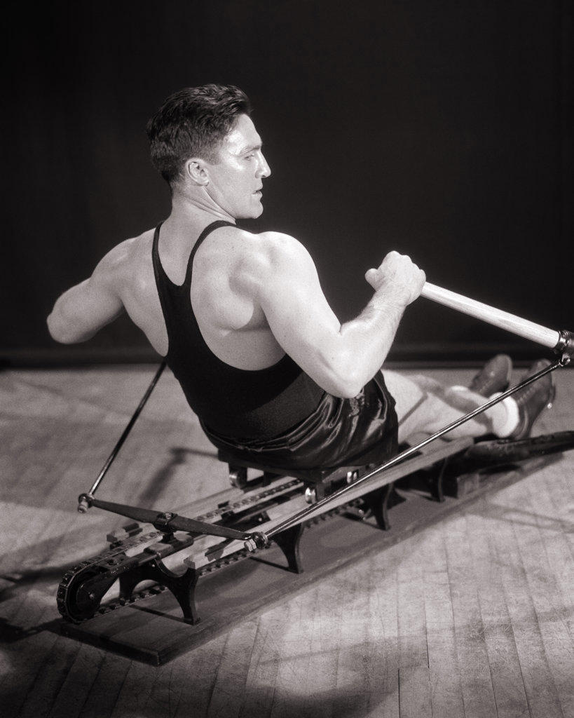 1930s MAN MUSCULAR ATHLETE CONCENTRATED FOCUSED FACIAL EXPRESSION EXERCISING WORKING OUT ON GYMNASIUM STATIONARY ROWING MACHINE