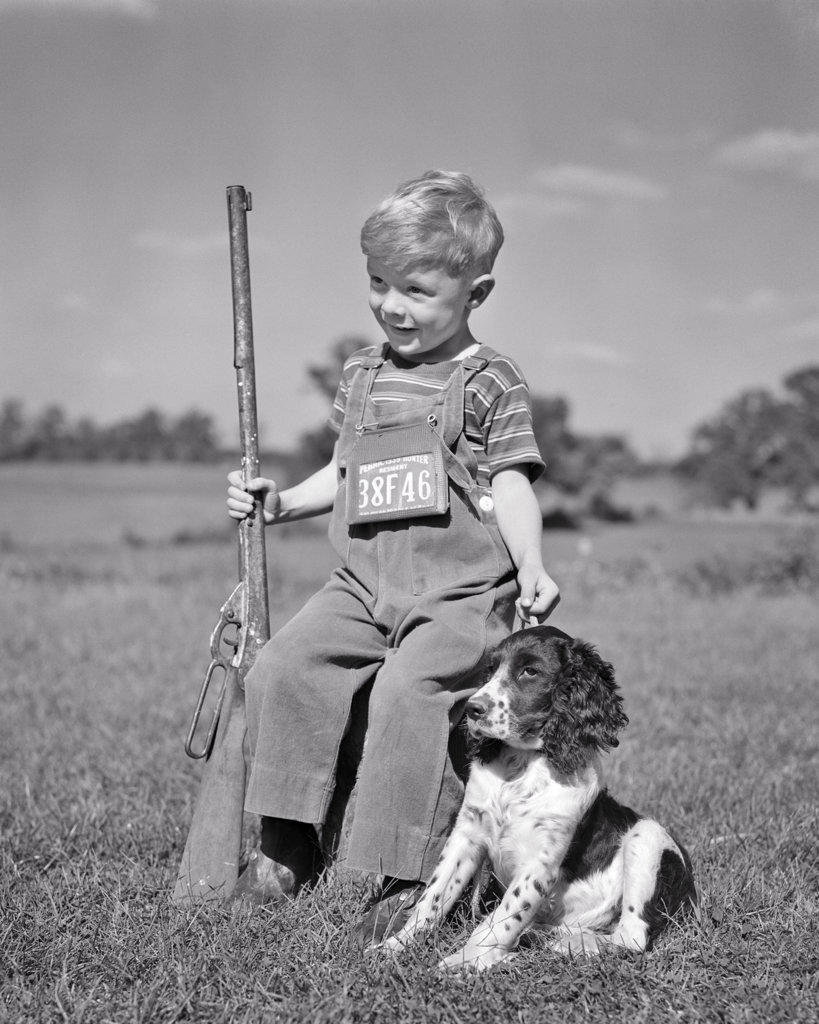 1940s SMILING BOY WEARING OVERALLS WITH HUNTING LICENSE AROUND HIS NECK SEATED WITH BB GUN AND SPRINGER SPANIEL DOG ALONG SIDE