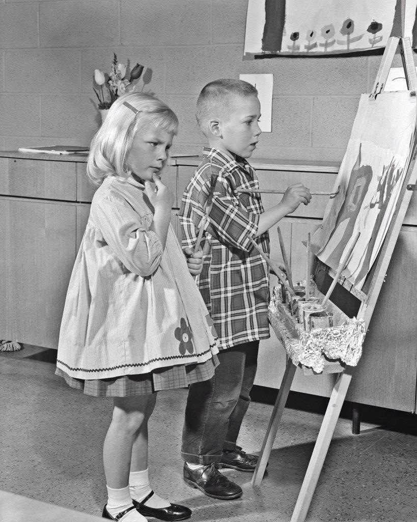 1960s CRITICAL YOUNG BLOND GIRL WATCHING BOY PAINTING ON EASEL ART CLASS AN ARTIST AND A CRITIC BOTH WEARING SMOCKS