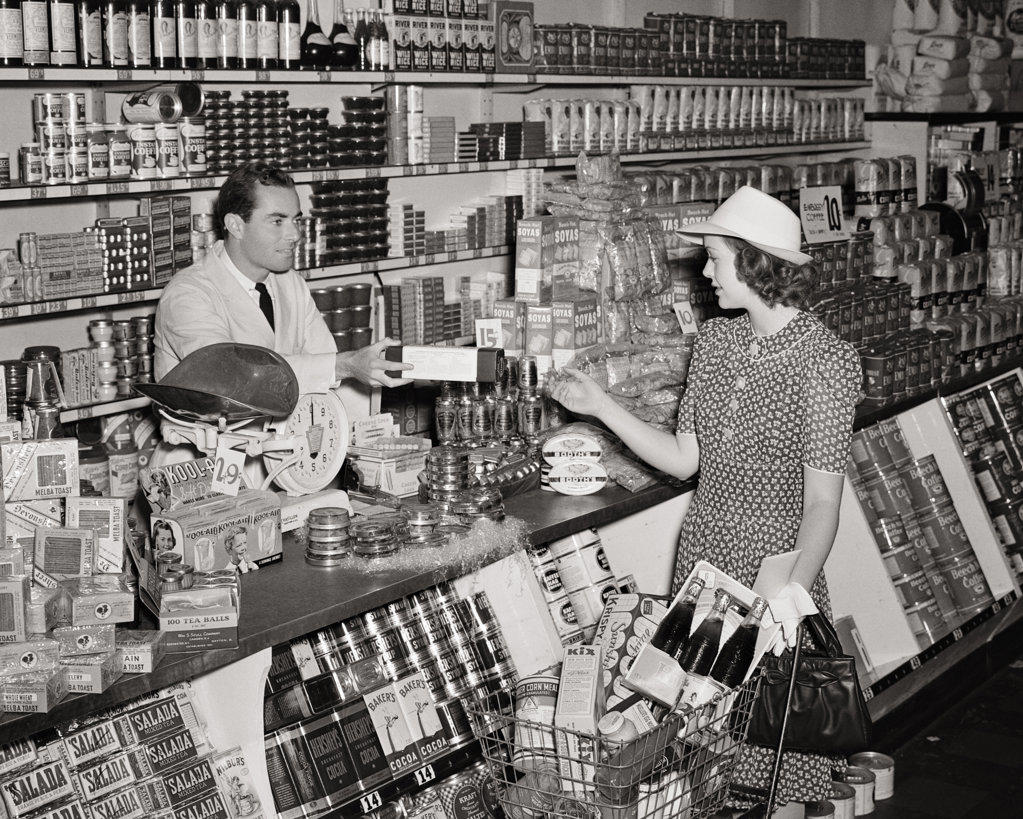 1940s WOMAN SHOPPING IN GROCERY STORE PUSHING SHOPPING CART ACCEPTING A PACKAGE FROM CLERK MAN STANDING BEHIND COUNTER