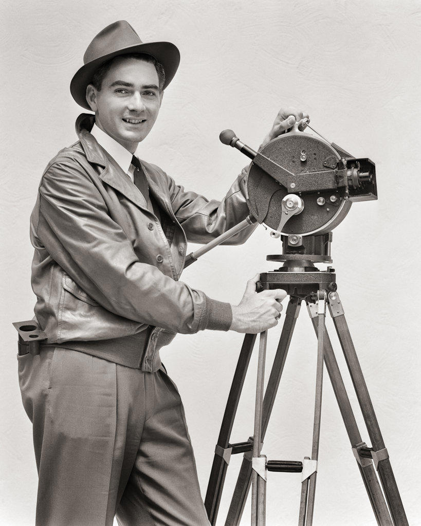 1930s SMILING MAN CINEMATOGRAPHER OPERATING AKELEY HAND CRANK MOVIE CAMERA LOOKING AT CAMERA WEARING LEATHER JACKET FEDORA HAT