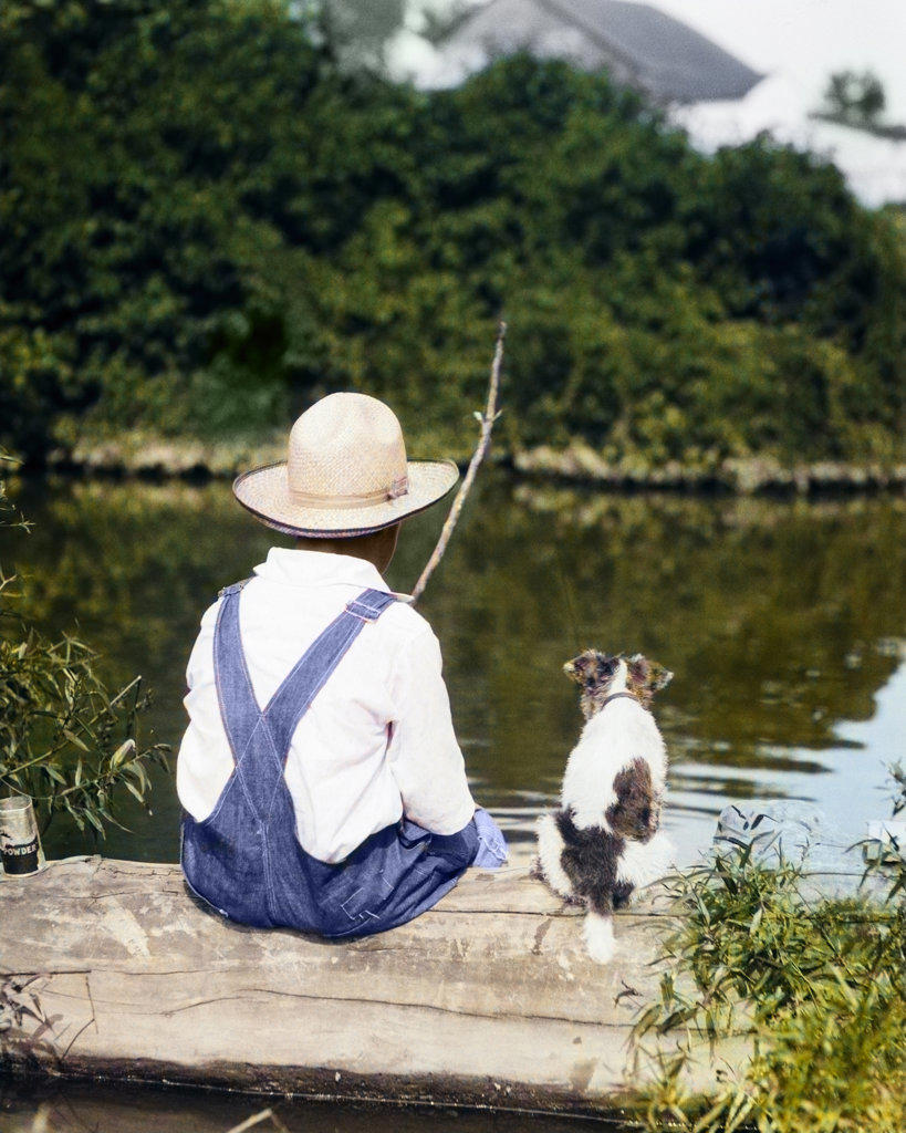 1920s 1930s FARM BOY WEARING STRAW HAT AND OVERALLS SITTING ON LOG WITH SPOTTED DOG FISHING IN POND