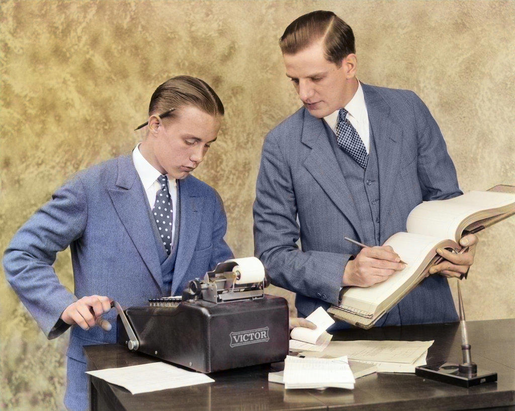 1920s BOOKKEEPER AND YOUNG ASSISTANT IN OFFICE USING LEDGER BOOK AND MANUAL ADDING MACHINE
