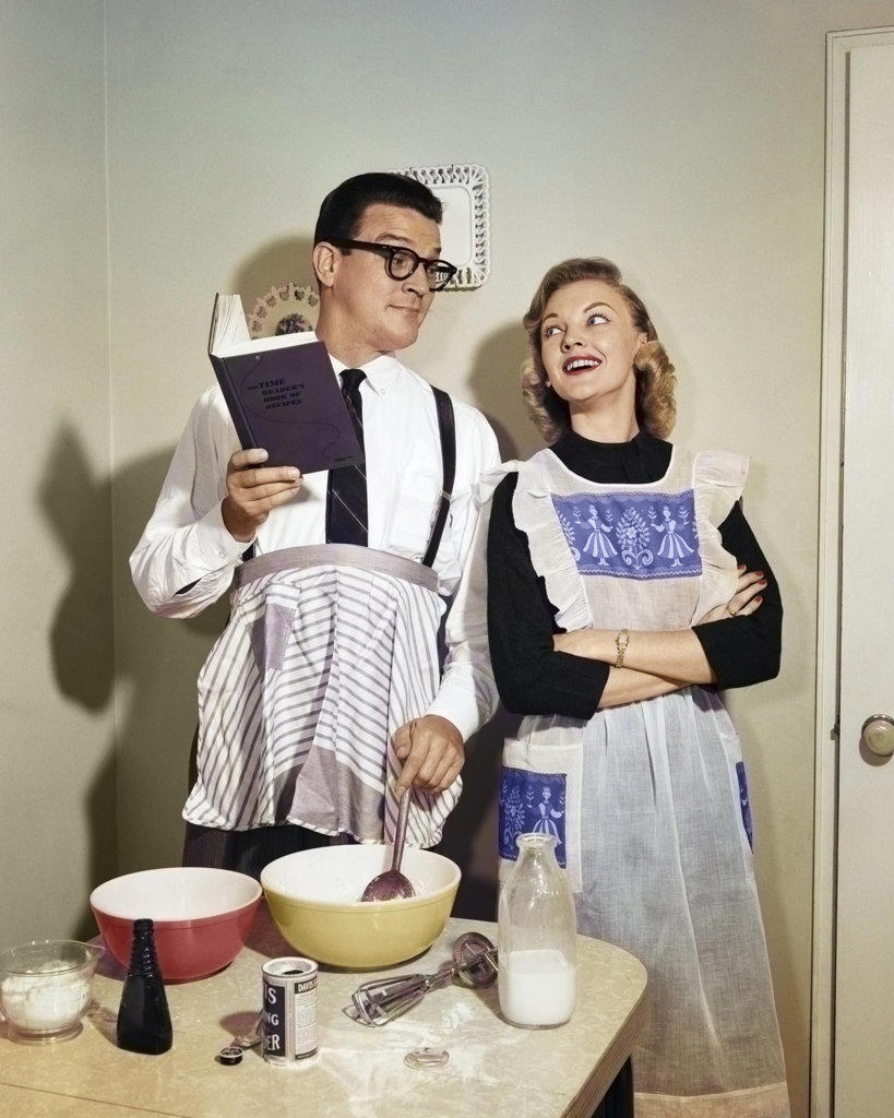 1950s SMILING COUPLE IN KITCHEN WITH HUSBAND LEARNING TO COOK FROM A BOOK WHILE WIFE WATCHES BOTH WEARING APRONS