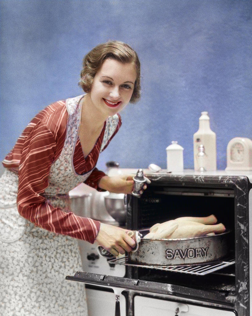 1930s SMILING HOUSEWIFE IN APRON LOOKING AT CAMERA SALTING SEASONING TURKEY IN SAVORY ROASTING PAN GOING INTO OVEN