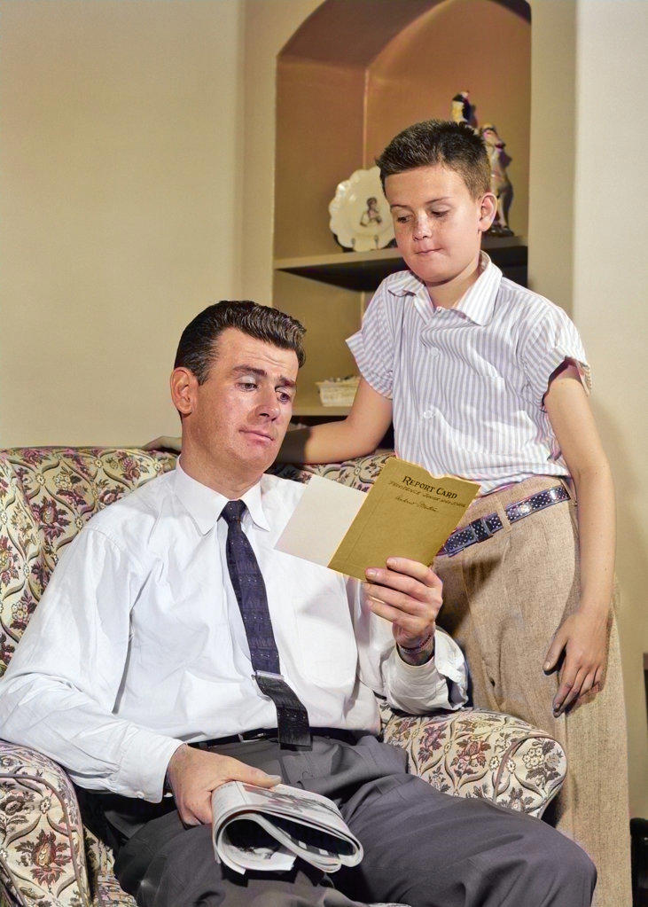 1950s WORRIED BOY STANDING LOOKING DOWN AT SKEPTICAL FATHER REVIEWING HIS REPORT CARD