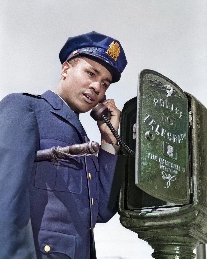 1960s AFRICAN AMERICAN POLICE OFFICER MAKING CALL AT POLICE BOX TELEPHONE NIGHT STICK TUCKED UNDER HIS ARM