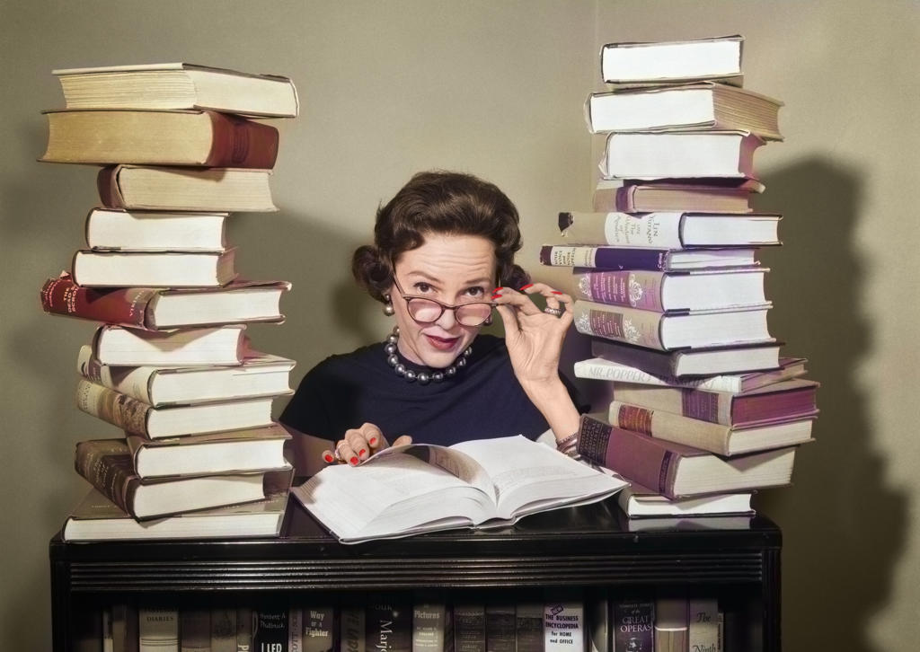1950s WOMAN LIBRARIAN SITTING BETWEEN STACKS OF BOOKS PULLING DOWN GLASSES TO LOOK OVER RIMS LOOKING AT CAMERA