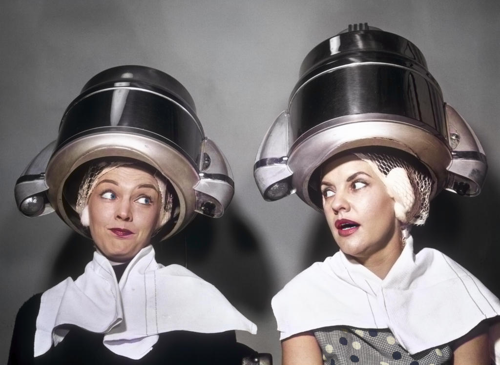 1950s TWO WOMEN GOSSIPING SITTING TOGETHER UNDER HAIR DRYERS IN HAIRDRESSER SALON