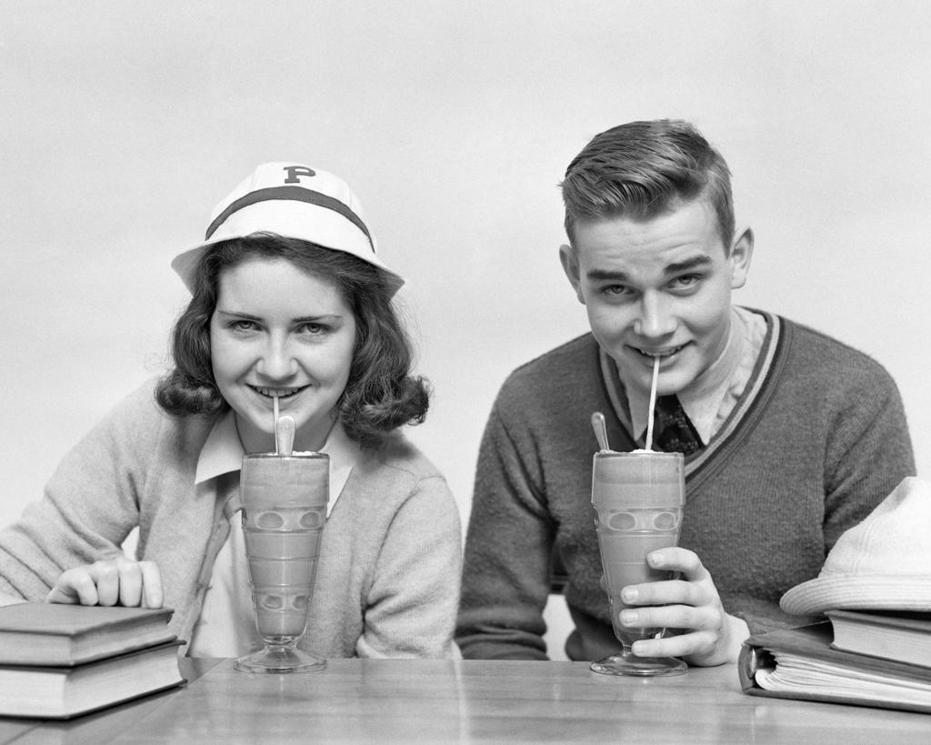 1940s TEENAGE BOY AND GIRL DRINKING MILKSHAKES TOGETHER LOOKING AT CAMERA