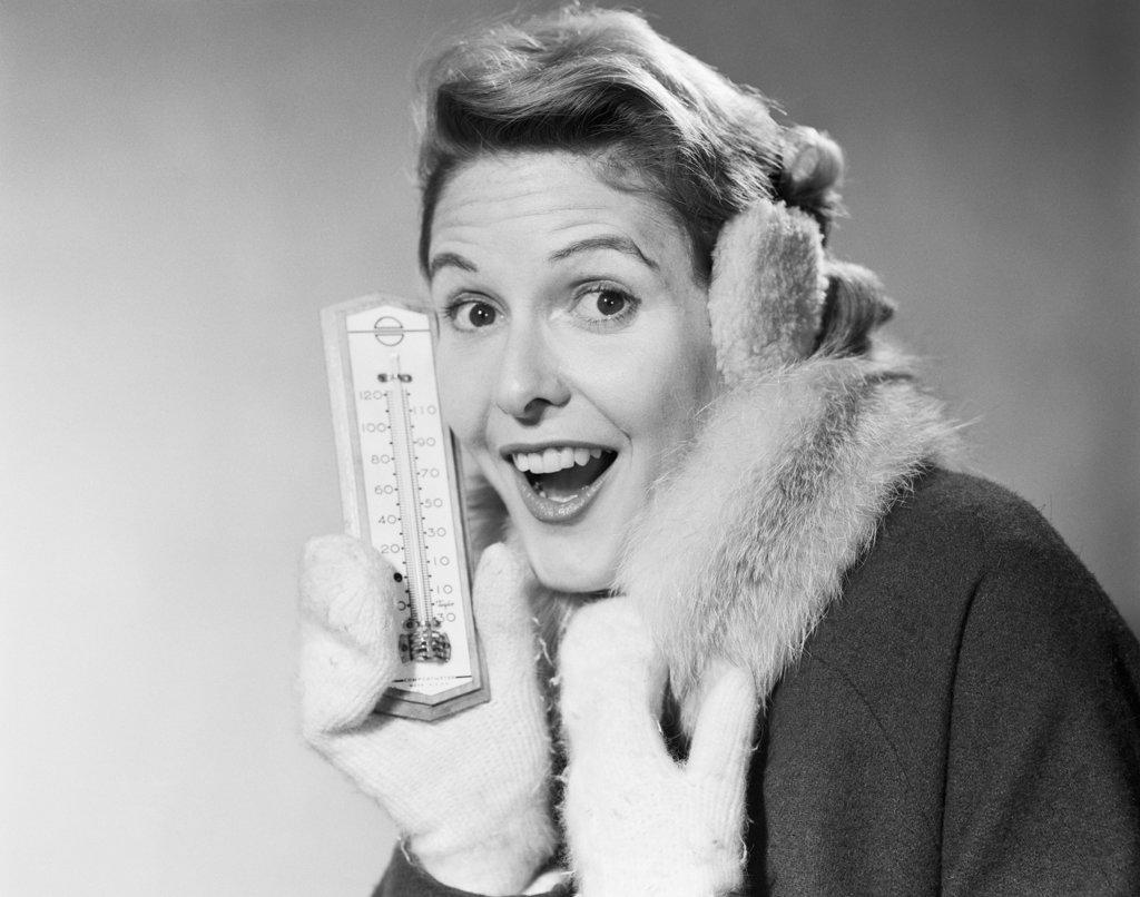 1950s SMILING WOMAN LOOKING AT CAMERA IN WINTER COLD HOLDING THERMOMETER WEARING COAT MITTENS EARMUFFS 
