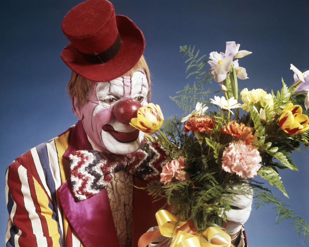 1970s PORTRAIT HAPPY CIRCUS CLOWN WEARING TINY RED TOP HAT STRIPED COSTUME BIG BOW TIE SMELLING BOUQUET OF FLOWERS