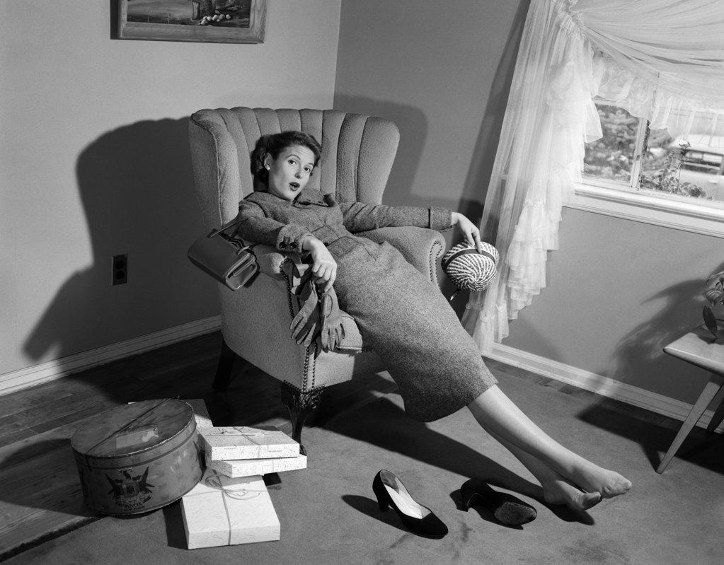 1950s WOMAN EXHAUSTED FROM SHOPPING SLUMPING IN LIVING ROOM CHAIR SHOES OFF PACKAGES EVERYWHERE LOOKING AT CAMERA 