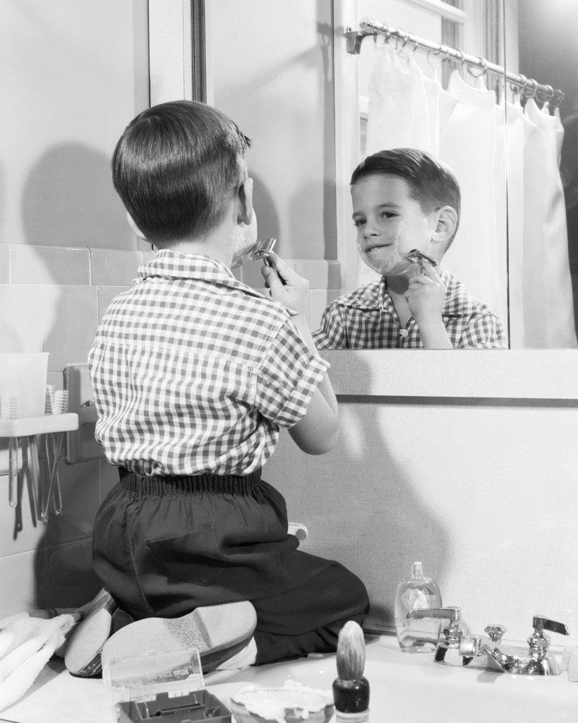 1950s BOY KNEELING BATHROOM SINK TRYING TO SHAVE HIS BEARD LIKE FATHER LIKE SON