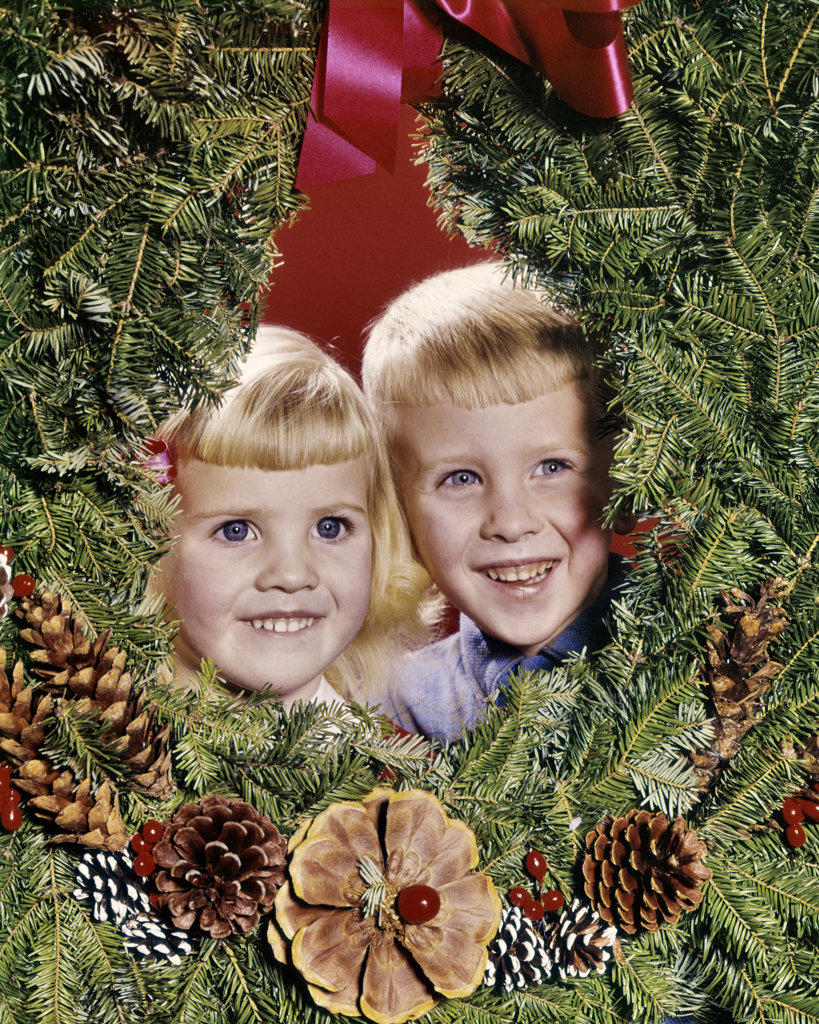 1960s SMILING BLONDE BOY AND GIRL BROTHER AND SISTER PEEKING OUT FROM CENTER OF CHRISTMAS WREATH