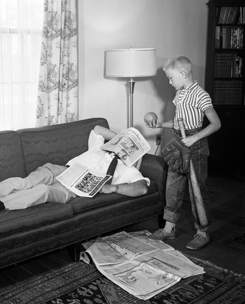 1950S Father Lying On A Sofa With Newspaper Over His Head While Son Is Standing Over Him With Bat Ball And Baseball Glove