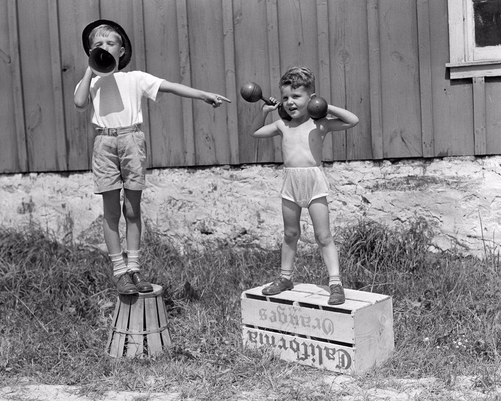 1930S 1940S Boys Playing Carnival Strongman One Lifting Dumbbells Other Announcing Act Through Megaphone