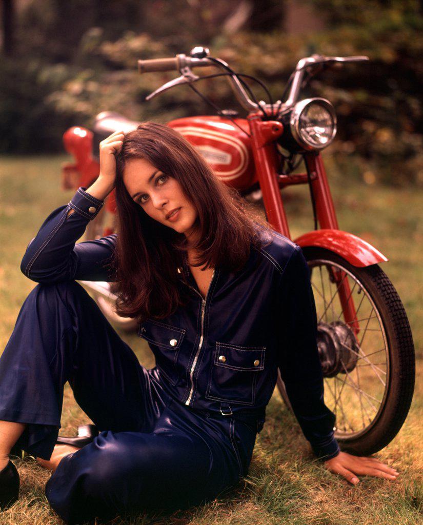 1970S Sexy Woman Long Brunette Hair Blue Leather Jumpsuit Sitting On Grass With Red Motorbike