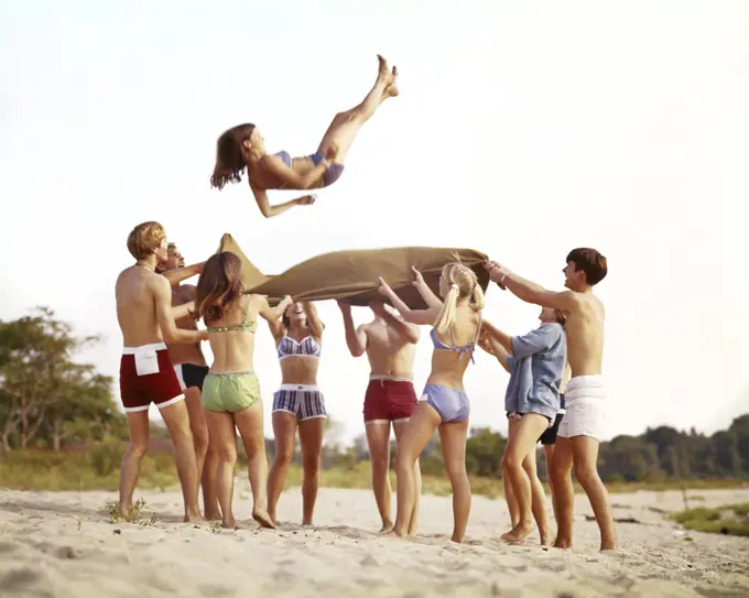 1960s 1970s BEACH GAMES GROUP OF TEENAGE BOYS AND GIRLS WEARING BATHING SUITS BEACH BLANKET TOSSING ONE GIRL INTO THE AIR