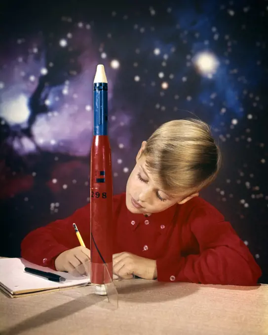 1960s BOY WITH MODEL ROCKET WRITING ON NOTE PAD WITH PENCIL STAR GALAXY IN BACKGROUND