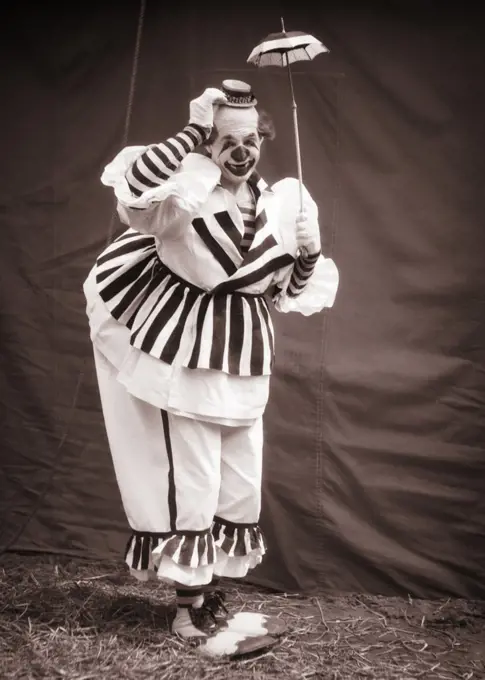 1930s BIG CLOWN UNDER TINY UMBRELLA TIPPING HIS SMALL HAT LOOKING AT CAMERA WEARING CLOWN SHOES OVERSTUFFED STRIPED COSTUME