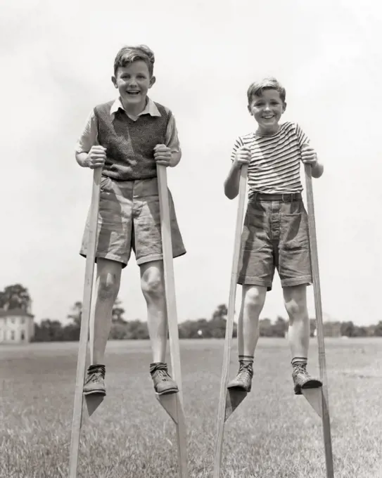 1930s TWO SMILING BOYS ON WOODEN HOMEMADE STILTS WEARING SHORTS BALANCING WALKING TOWARDS LOOKING AT CAMERA SUMMERTIME