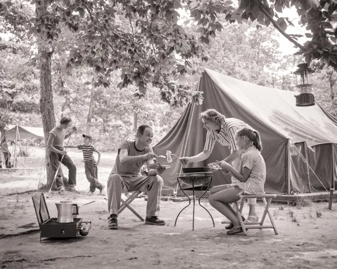 1950s 1960s FAMILY OF 5 TENT CAMPSITE MOM COOKING BREAKFAST SERVING DAUGHTER AND DAD TWO BOYS LEANING ON TREE WITH FISHING ROD