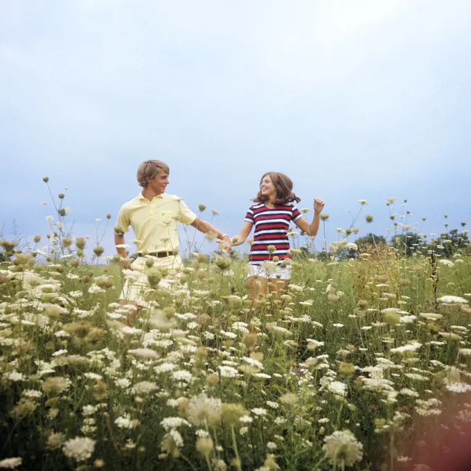 1970s TEENAGE COUPLE HOLDING HANDS RUNNING THROUGH A FIELD OF QUEEN ANNE’S LACE SMILING WEARING SUMMER CLOTHES