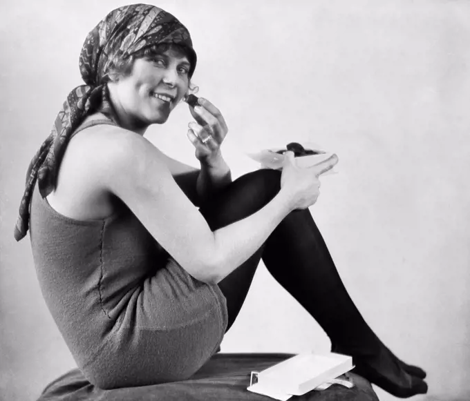 1920S Woman In Old Fashioned Bathing Suit With Scarf On Head Seated With Knees To Chest Eating Chocolates