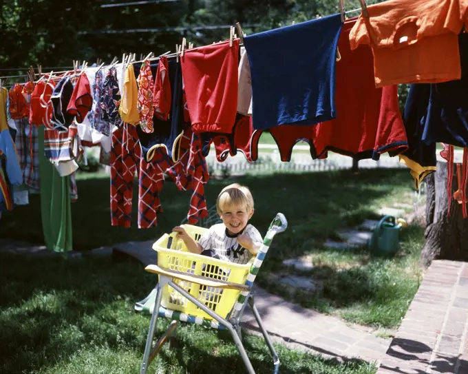 1970s LAUGHING BLOND BOY LOOKING AT CAMERA SITTING IN YELLOW LAUNDRY BASKET UNDER CLOTHESLINE FULL OF COLORFUL WASHED CLOTHES