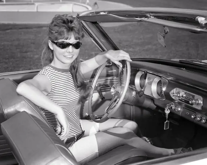 1960s SMILING  PRETTY TEEN GIRL WEARING SUNGLASSES LOOKING AT CAMERA SITTING AT STEERING WHEEL IN CONVERTIBLE CAR DRIVER’S SEAT