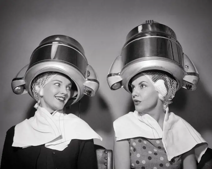 1950s TWO WOMEN SITTING UNDER BEAUTY SALON HAIR DRYERS WEARING HAIRNETS TOWELS TALKING GOSSIP 