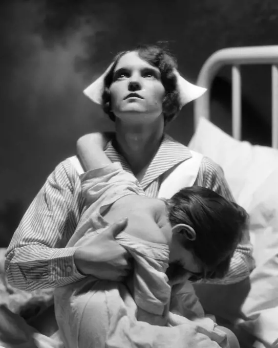 1920s 1930s COMPASSIONATE PRAYING NURSE HOLDING A SICK CHILD IN HOSPITAL BED PORTRAIT DRAMATIC LIGHTING