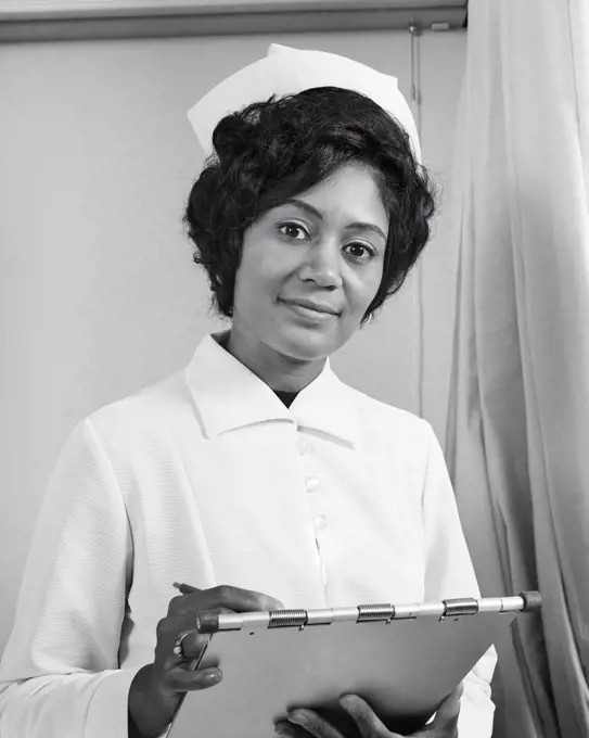 1970s SERIOUS AFRICAN-AMERICAN WOMAN NURSE IN UNIFORM LOOKING AT CAMERA ENTERING PATIENT INFORMATION IN HOSPITAL CHART