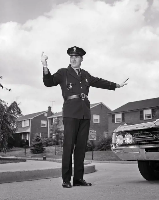 1960s SINGLE SMILING UNIFORMED POLICEMAN ARMS HANDS EXTENDED STANDING AT INTERSECTION DIRECTING SUBURBAN AUTOMOBILE TRAFFIC 