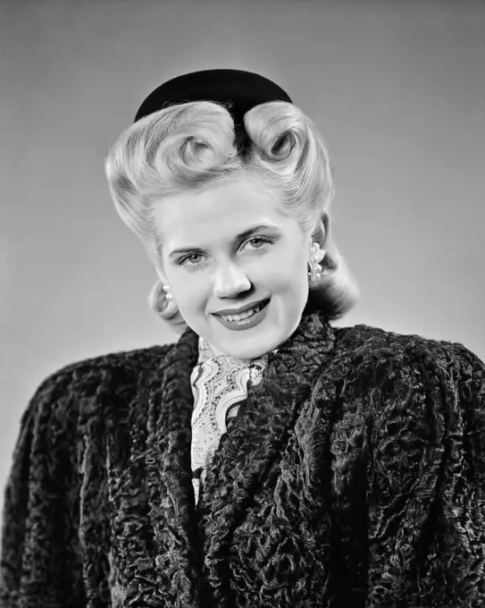 1940s ELEGANT WELL-TO-DO BLOND WOMAN WITH VERY BIG VICTORY ROLLS HAIR STYLE LOOKING AT CAMERA WEARING BLACK PERSIAN LAMB COAT