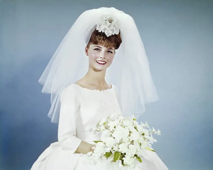 1960s SMILING BRUNETTE BRIDE PORTRAIT IN SIMPLE WHITE WEDDING GOWN HOLDING WHITE FLOWER BOUQUET WEARING VEIL LOOKING AT CAMERA