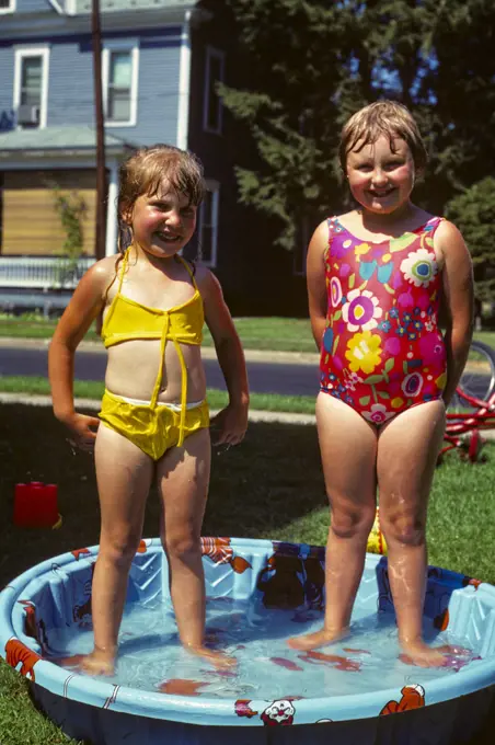 1970s TWO SMILING GIRLS WEARING MESSY WET BATHING SUITS YELLOW AND RED STANDING IN KIDDIE POOL IN BACKYARD LOOKING AT CAMERA 