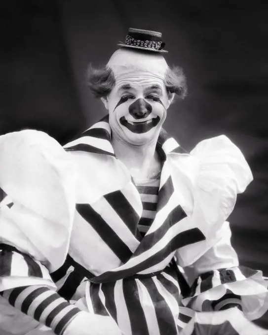 1930s SMILING BIG TOP CIRCUS WHITEFACE CLOWN LOOKING AT CAMERA WEARING TINY HAT STRIPED OVERSIZE COSTUME
