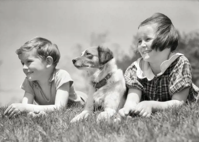 1930s SMILING BOY AND GIRL A BROTHER AND SISTER LYING TOGETHER IN THE GRASS WITH THEIR PET DOG BETWEEN