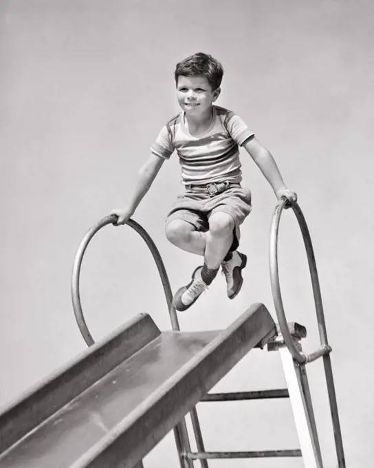 1940s BOY AT TOP OF PLAYGROUND SLIDE PUSHING UP ON HAND RAILS CROSSING HIS LEGS FUNNY FACIAL EXPRESSION 