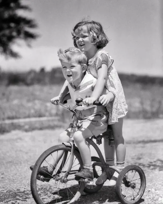 1930s BOY AND GIRL BROTHER SISTER RIDING TANDEM ON TRICYCLE GIRL STEERING ON BACK OF TRIKE BOY PEDDLING LAUGHING HAVING FUN 